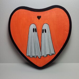A halloween-themed heart shaped plaque featuring two ghosts looking into eachother's eyes while standing before a small black heart.