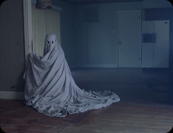 In this still from the movie 'A Ghost Story', the ghost looks towards the viewer while kneeling by an entryway, picking at a seam in the joinery/moulding.